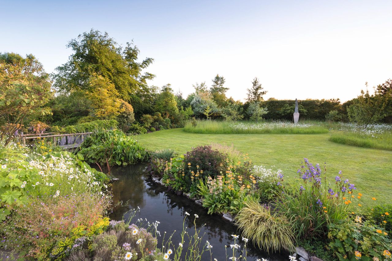 Iris sibirica, Primula bulleyana and Geum ‘Totally Tangerine’ by the stream in the garden at Midsummer House, Oxfordshire. Behind is the sculpture Ascent by Johannes von Stumm.