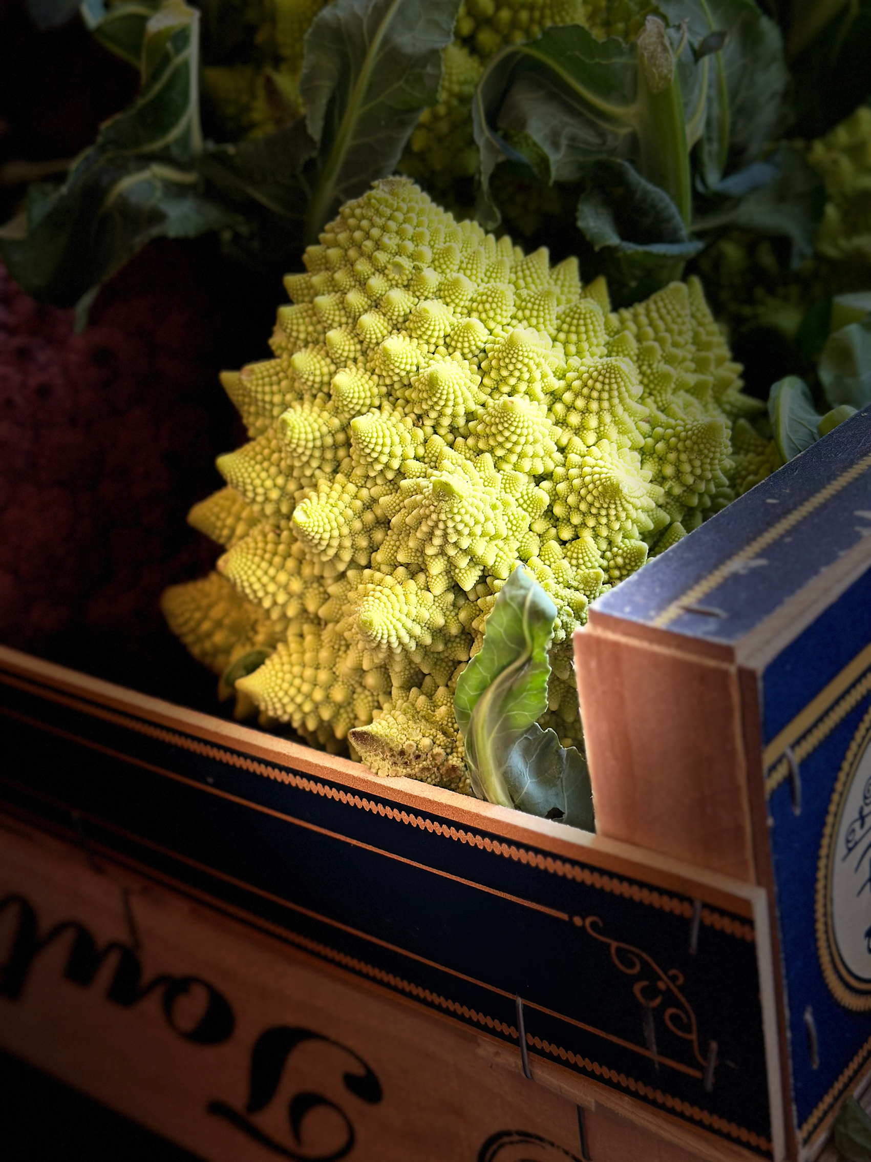 Close-up of a fresh Romanesco cauliflower with its distinctive fractal pattern, partially surrounded by green leaves in a wooden crate