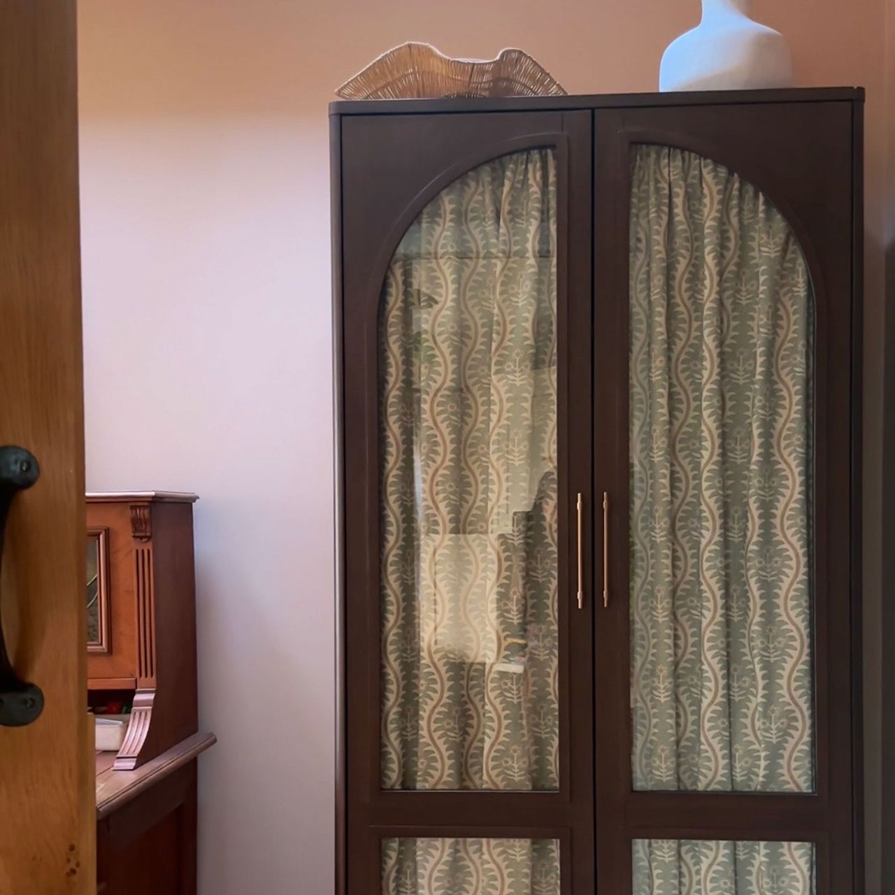 Pink room with wooden glass fronted cabinet with patterned curtains behind glass
