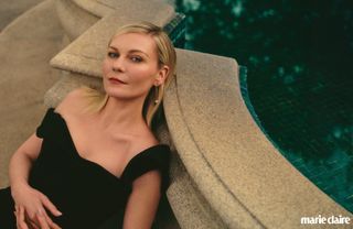 Kirsten Dunst leans against a pool wearing a black dress and gold earrings for with marie claire logo in bottom right corner for Marie Claire's Makers Issue 2024