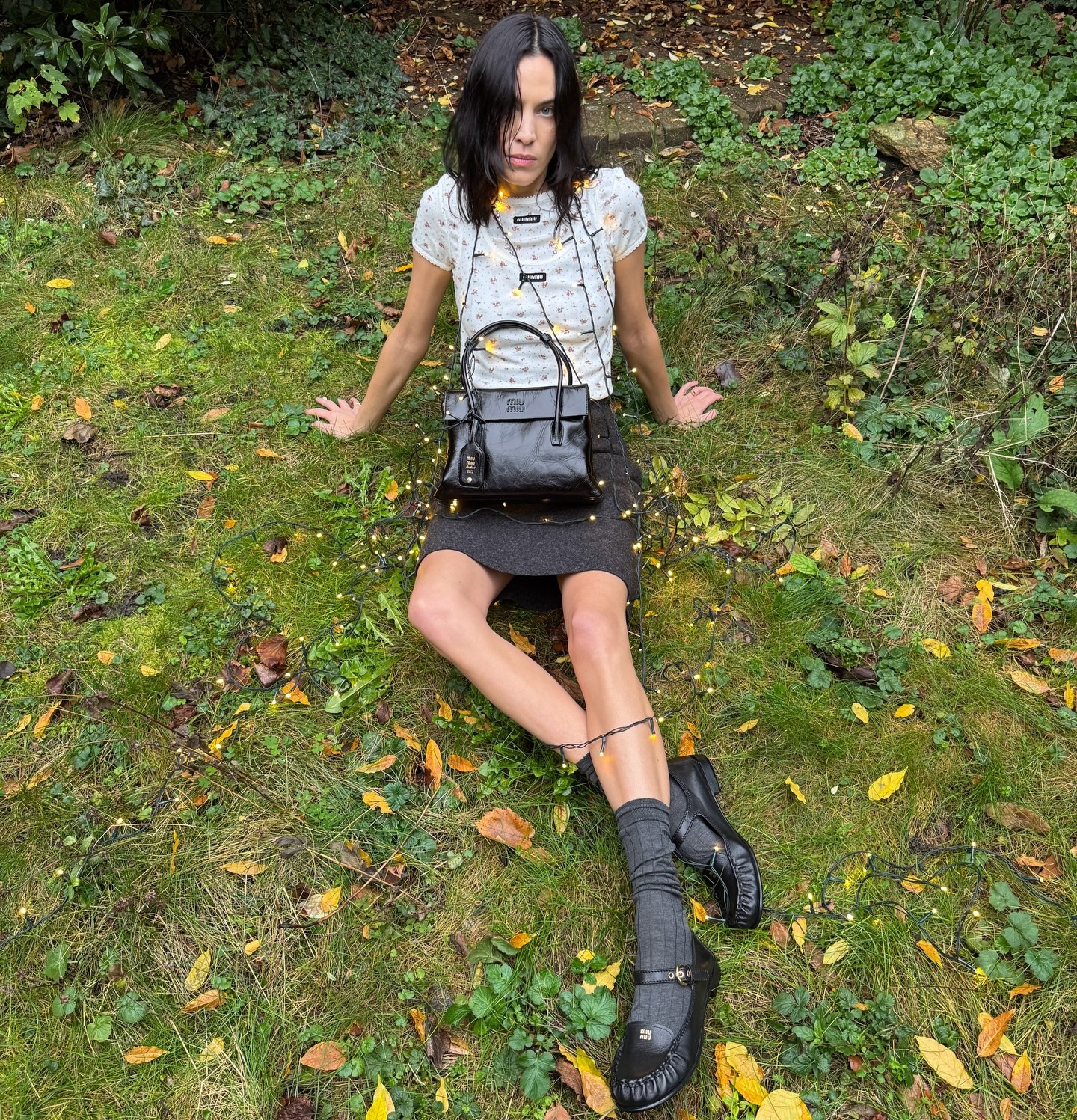 Alexa Chung sits in a garden wearing a layered top, a wool miniskirt and Miu Miu's Mary Jane loafers.