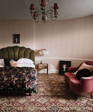Vintage style bedroom with red patterned carpet, pink patterned wallpaper, white ceiling, chandelier with fabric shades, bed with green scalloped headboard, deep red sofa with cushions