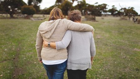 An adult daughter walks with her arm over her mom's shoulder.