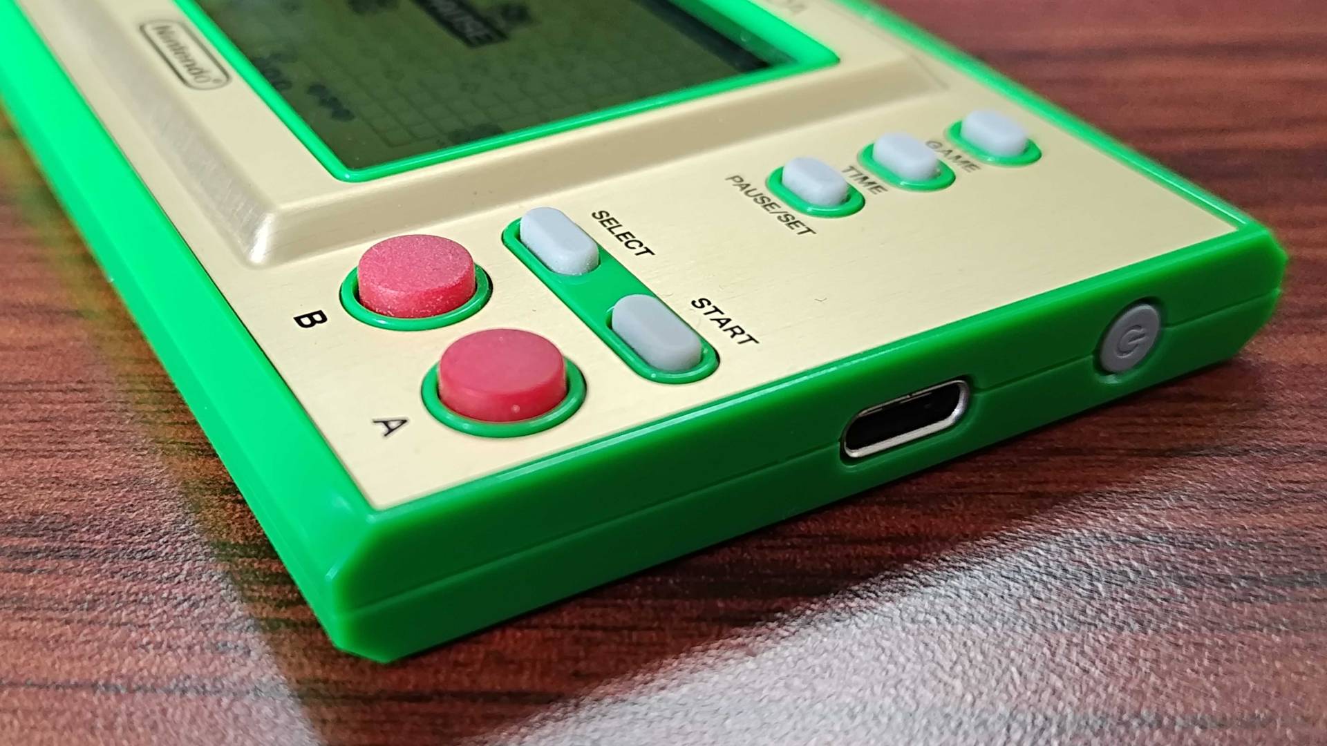 Close up of Zelda Game & Watch buttons, USB-C port, and power button with handheld sitting on woodgrain desk.