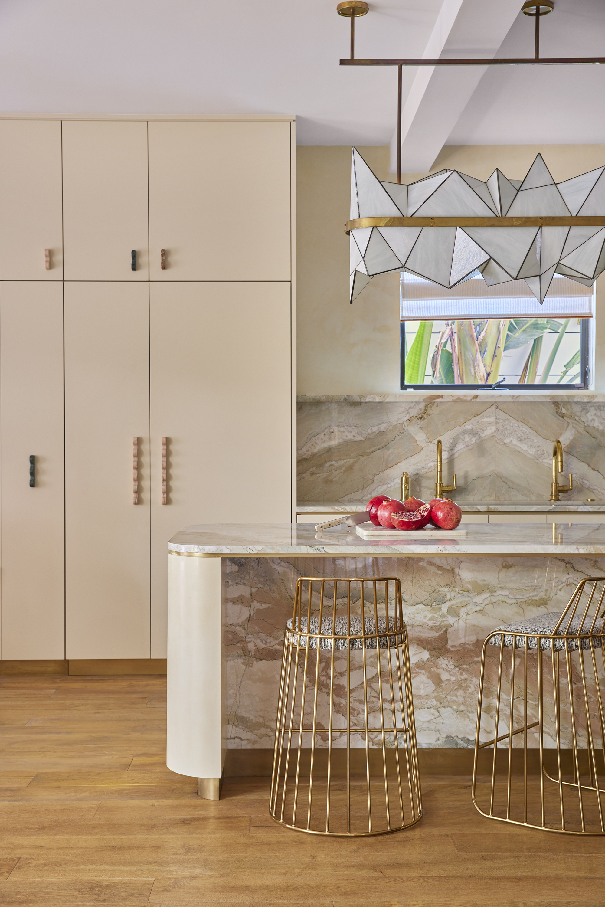 a beige kitchen with a marble island and backsplash, a floor to ceiling cabinet with detailed handles, and a geometric hanging pendant light