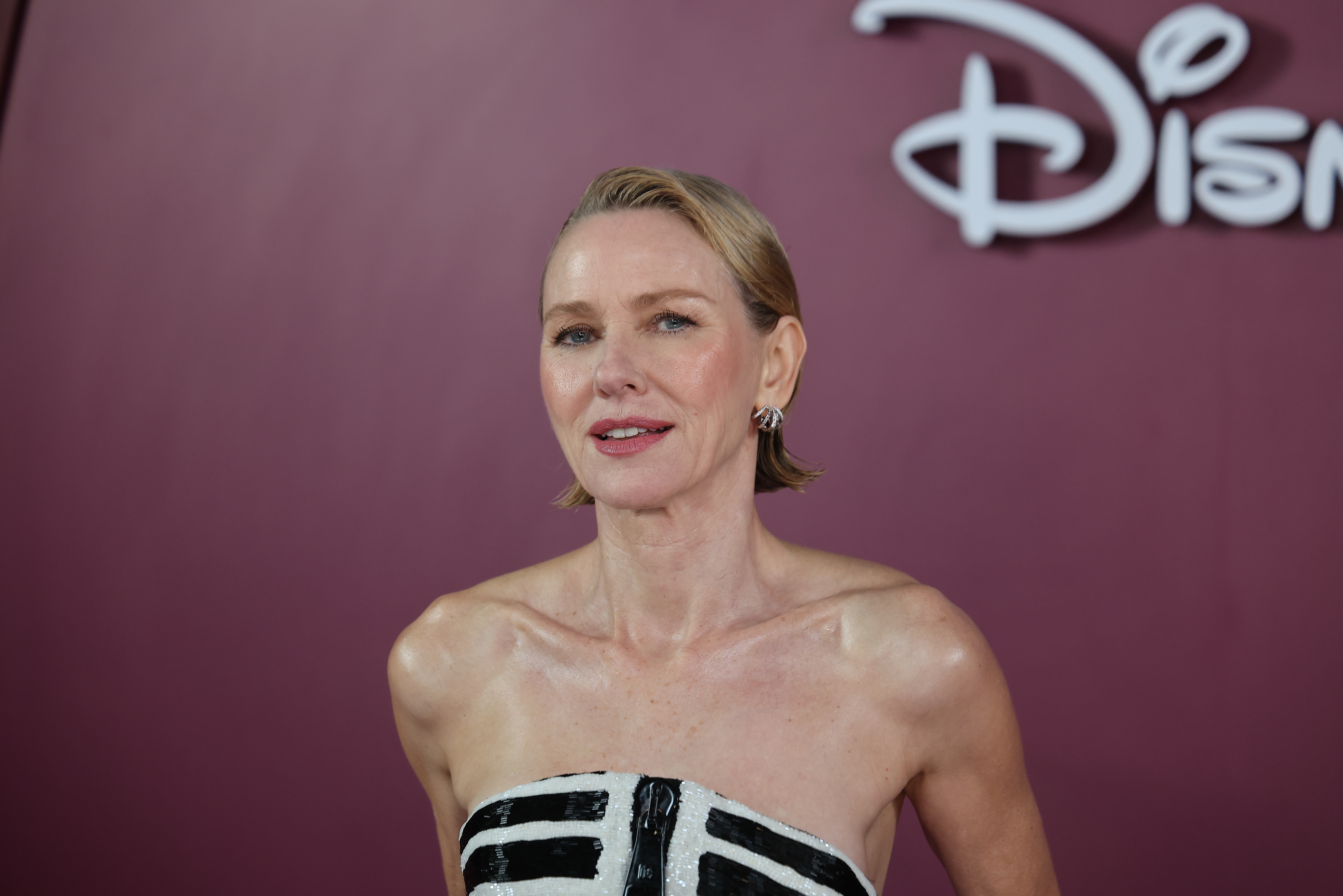 a photo of a white woman with short blonde hair posing in front of a mauve background at a red carpet premiere event