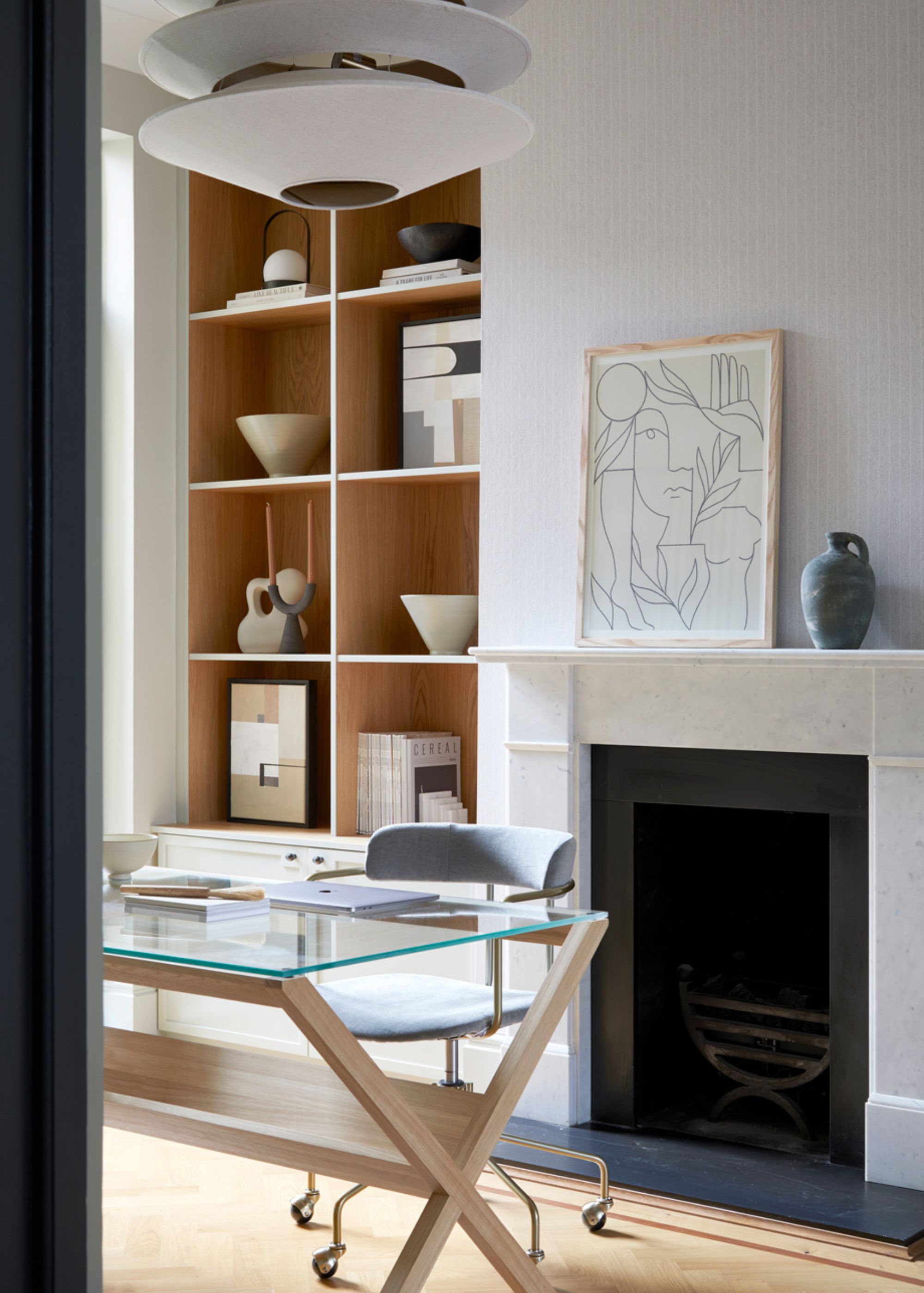 A modern home office with neutral walls, a white fireplace, a glass desk with a grey swivel chair, and built-in shelving in an alcove.