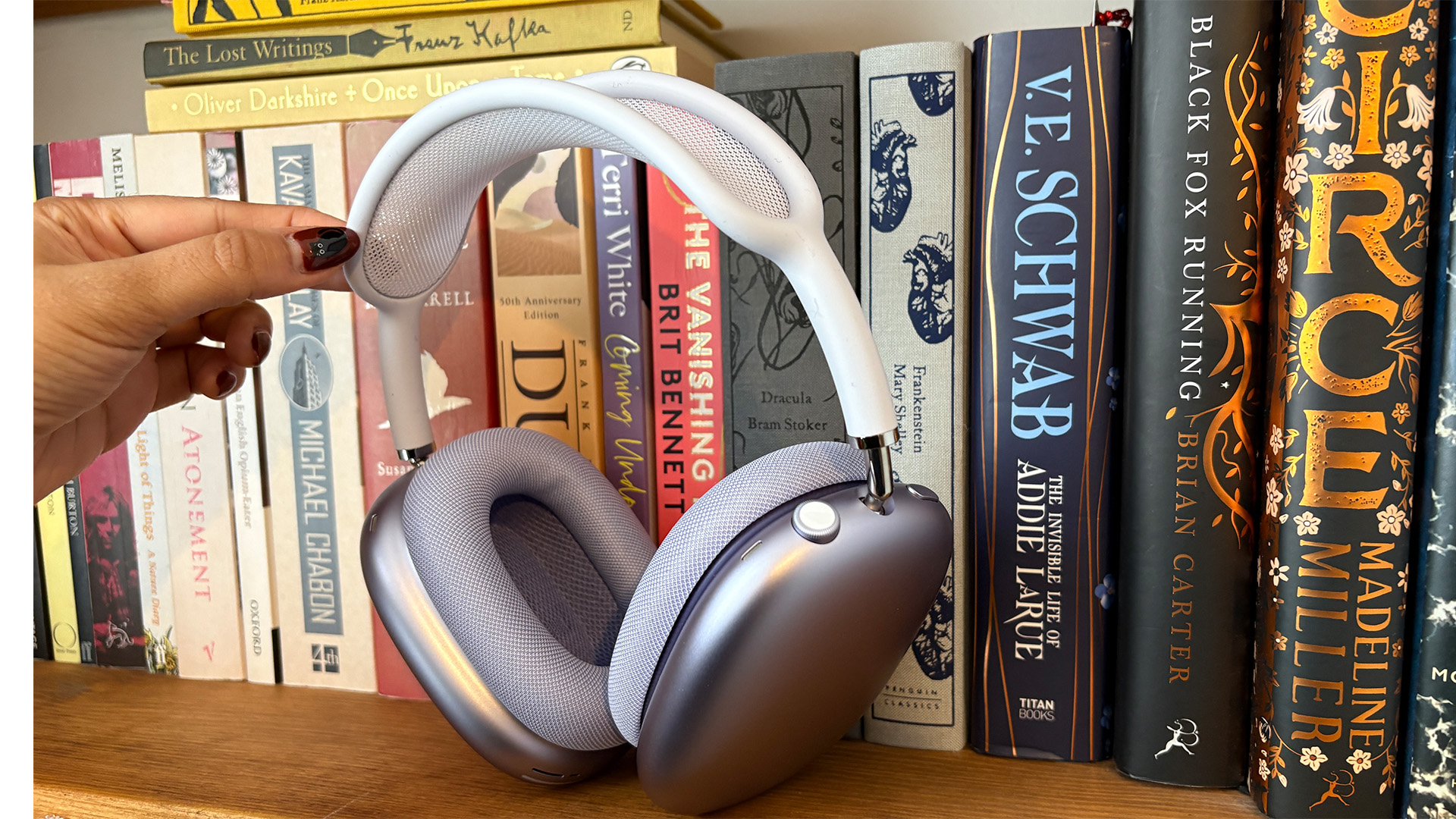 Apple AirPods Max 2 over-ear headphones on bookcase with hand holding them up by headband