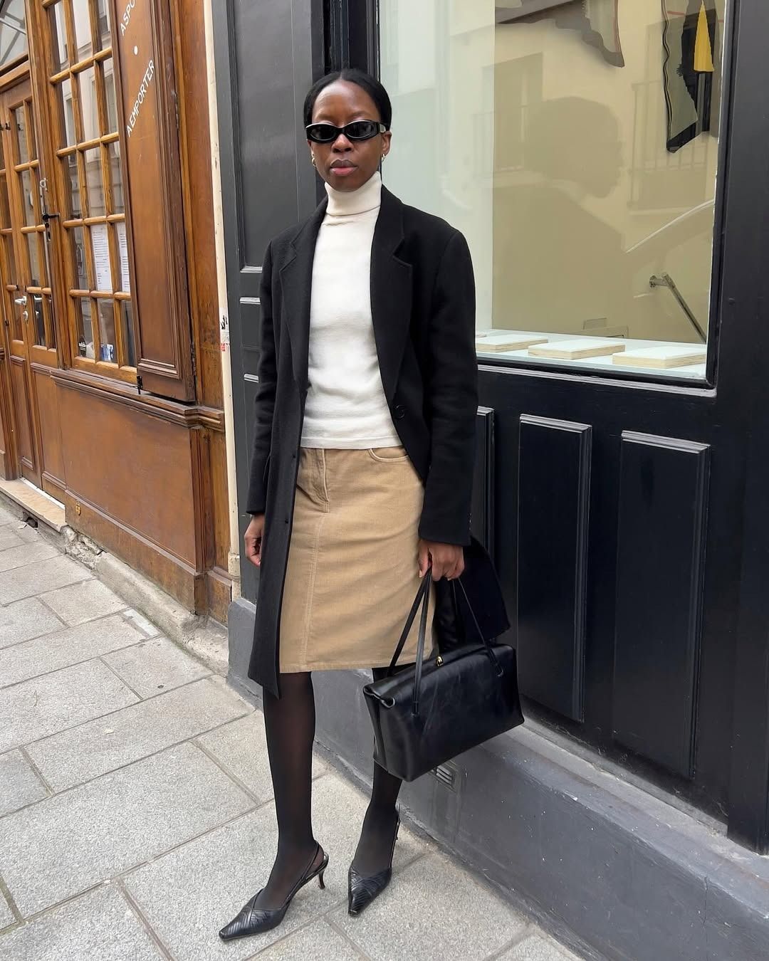 sylvie wears a black coat, white turtleneck, long black jacket, sheer black tights and black slingback heels.