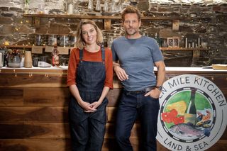 Martha (Sally Bretton) and Archie (Jamie Bamber) stand in front of the counter at Martha's café Ten Mile Kitchen. Martha has her hands clasped in front of her, and Archie is casually leaning on the counter.
