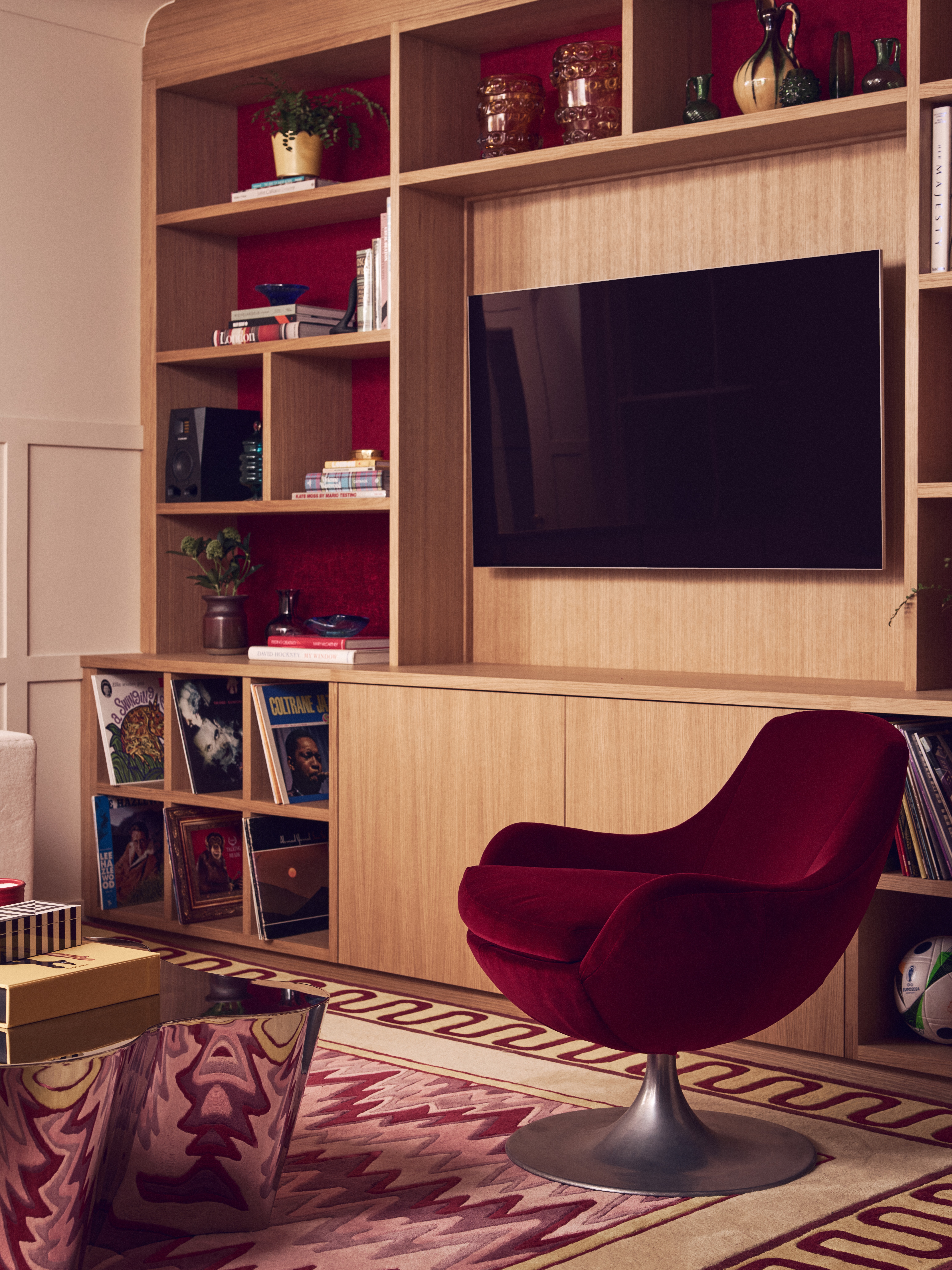 A plush deep-red velvet swivel chair sits in a modern room facing built-in light-oak shelving with a mounted TV, red accent backing, record storage, and a patterned area rug.