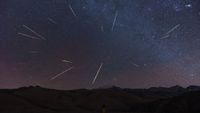 a dark mountainous landscape beneath a dark sky with many shooting stars