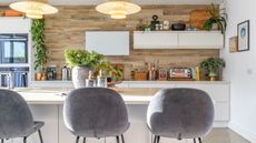 A bright kitchen with white cabinets and a wood effect back wall. A kitchen island with three large fabric barstools pulled up to it, plants on the counters, and two large pendant lights hanging above. 