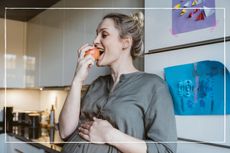 Pregnant woman eating an apple in her kitchen with kids artwork on the wall