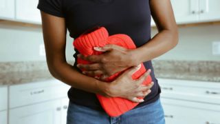 A woman wearing a black t-shirt holds a red water bottle against her to ease period pains