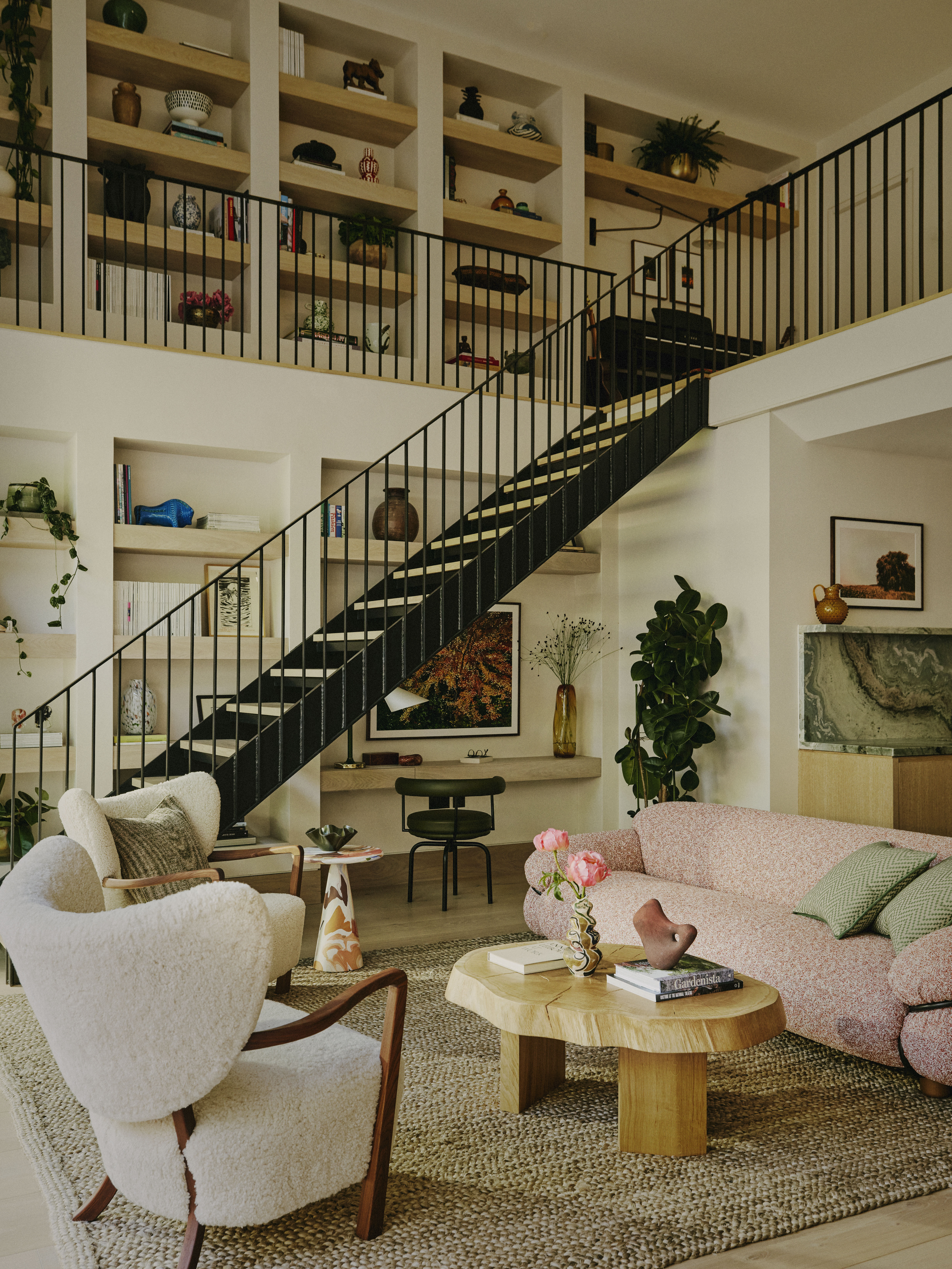 A living room with cream armchairs and pink sofas, styled around a chunky wooden coffee table, integrated shelving, and unique artwork, features a statement staircase in wrought iron and wood.