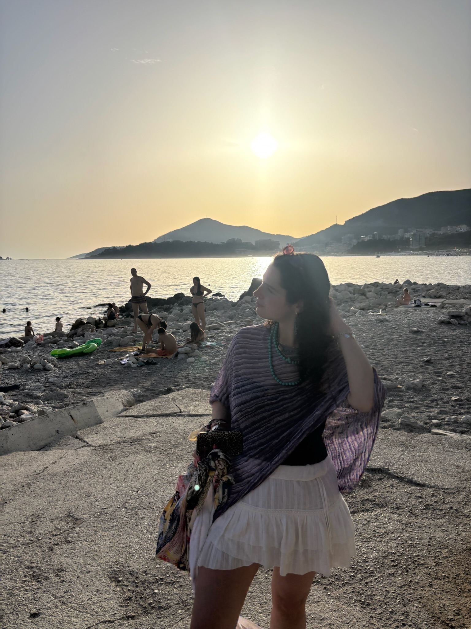 A young woman dressed in a white mini skirt and a wide purple top stands on a rocky beach at dusk.