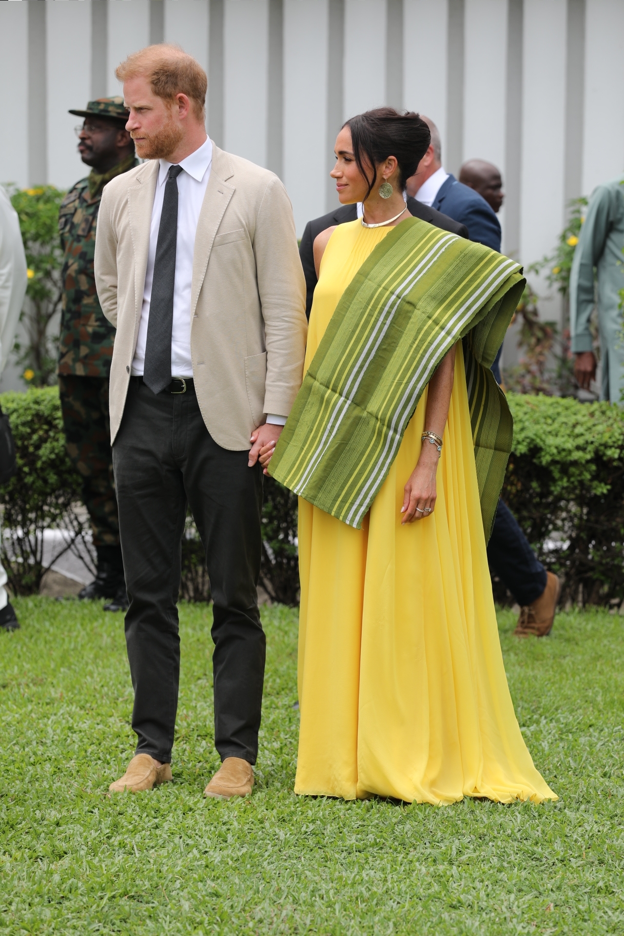 Prince Harry wearing a suit jacket and blue tie holding hands with Meghan Markle in a yellow dress and green scarf draped over her shoulder