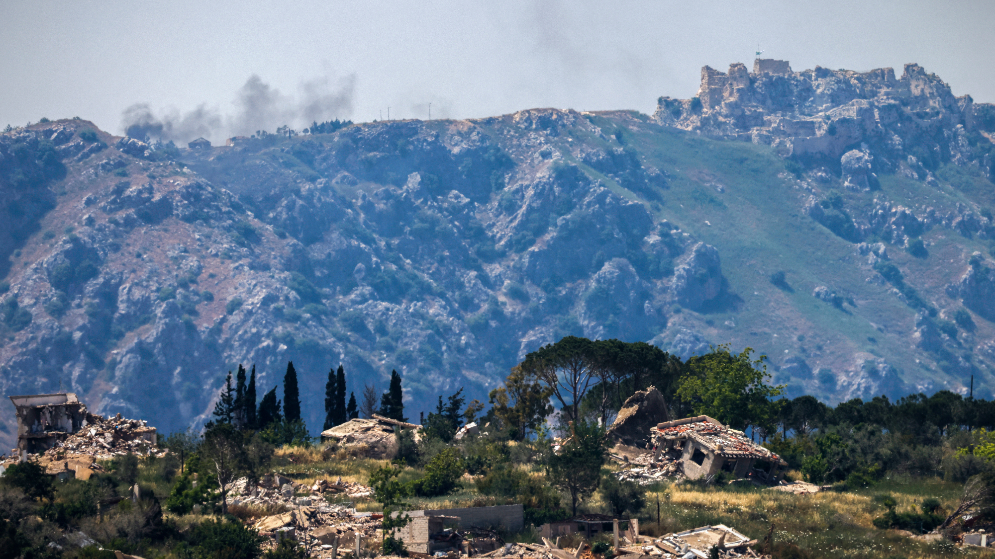 Destroyed buildings in the southern Lebanese village of Kfar Kila, with smoke rising in the distance behind the medieval Beaufort Castle, known locally as Qalaat al-Shaqif or Shaqif Arnoun, following an Israeli airstrike.