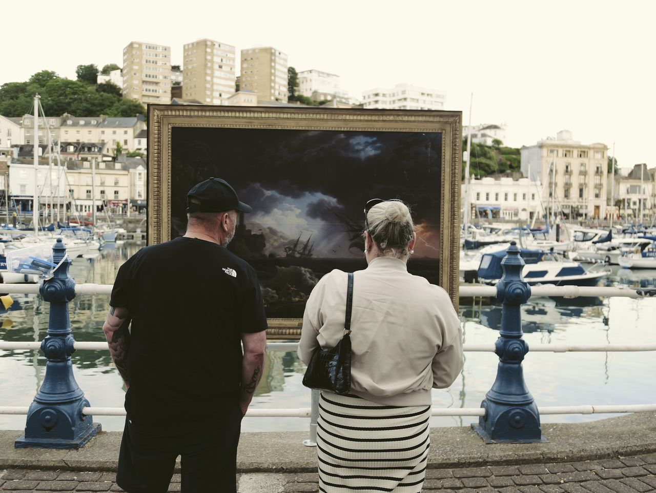 Sample image taken with the Panasonic Lumix L10; a couple looking at a classic painting which has been positioned outdoors in a seaside harbour