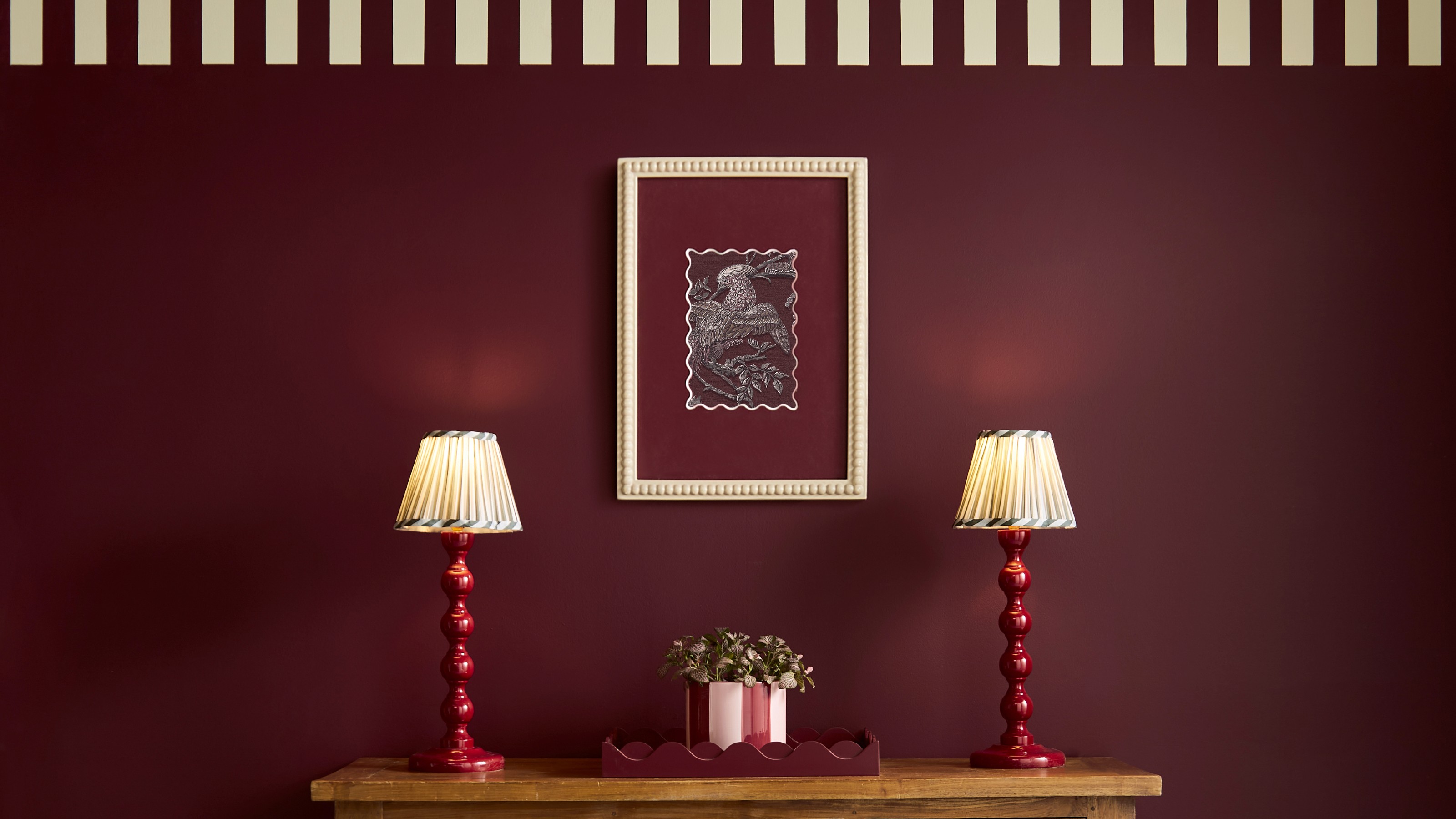 A wall painted in Graham &amp; Brown&#039;s Divine Damson with a striped trim and a console table with two red lamps