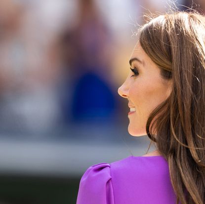 A shot of Kate Middleton from the back wearing a purple dress and looking to the side smiling 