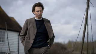 Tom Hiddleston in a brown suit, navy jumper and white dog collar by the shore as Will Ransome in The Essex Serpent
