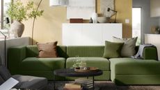 ikea living room with green L-shaped sofa, yellow wall, white cabinet behind, round coffee table, vase with branches on the windowsill, light leaning over sofa, and decor