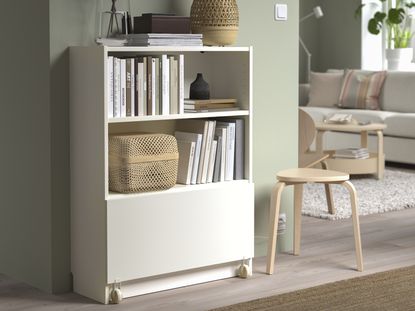 A living room with a small white bookcase against a wall with a pull out drawer with wheels at the bottom