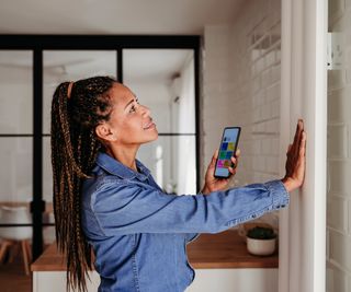 smiling woman with long dark hair in braids touching vertical radiator and holding phone in hand