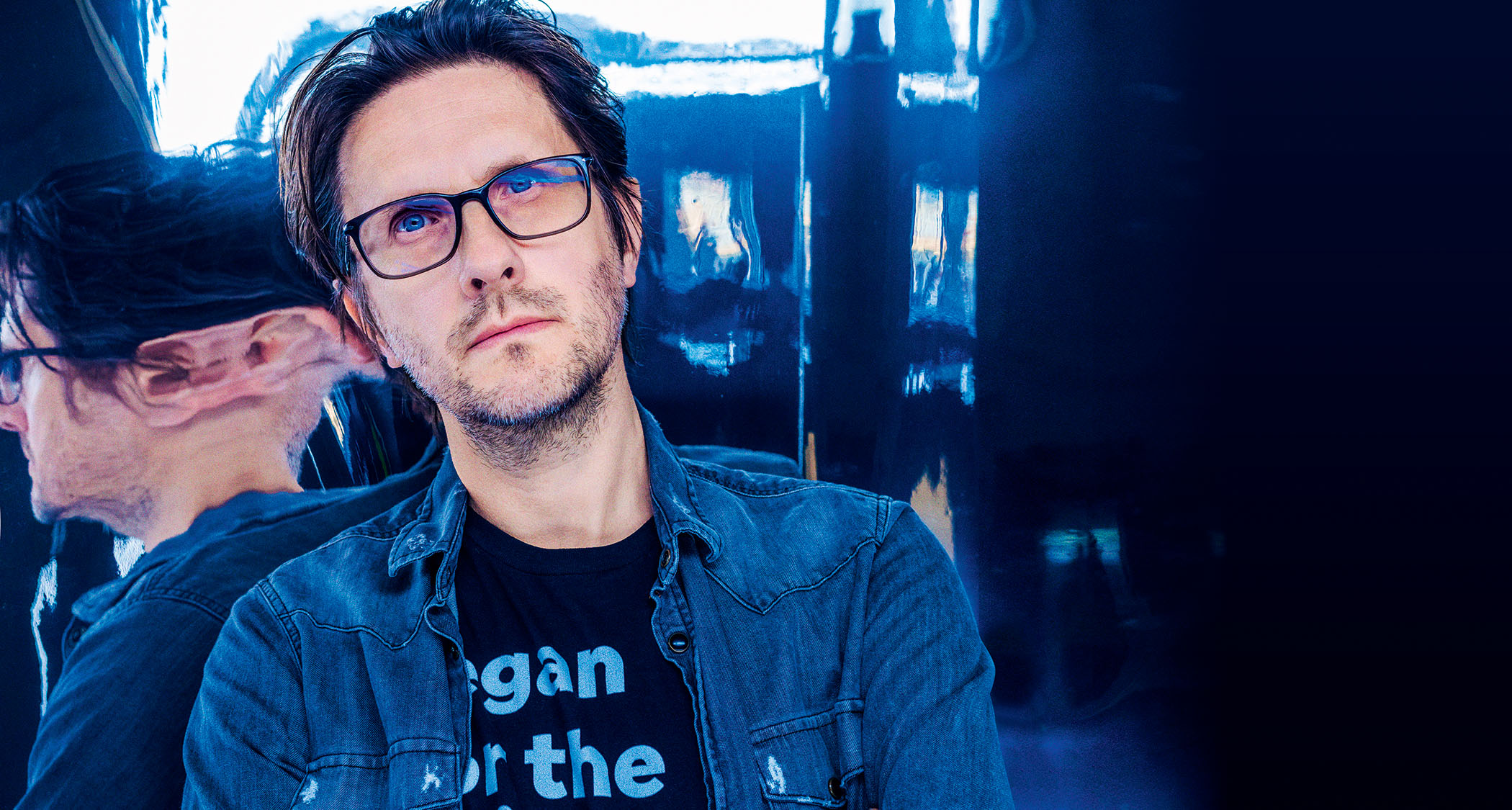 Steven Wilson wears a denim shit and a black T-shirt and is photographed against a distorted reflective surface.