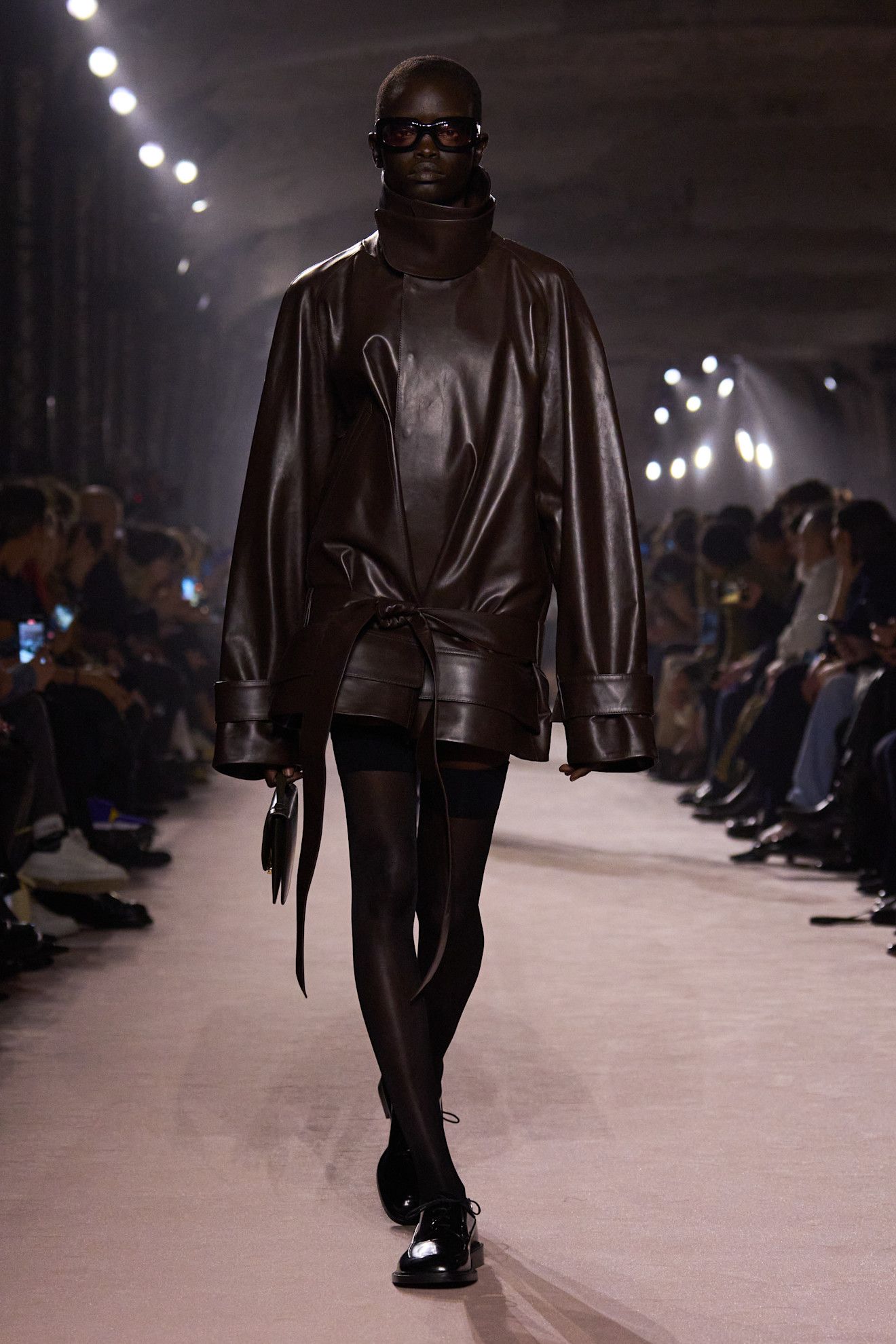 A model walking in the Victoria Beckham show wearing a brown leather funnel-neck jacket.