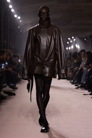 A model walking in the Victoria Beckham show wearing a brown leather funnel-neck jacket.