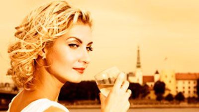 blond woman leaning on a fence in a field drinking a glass of wine