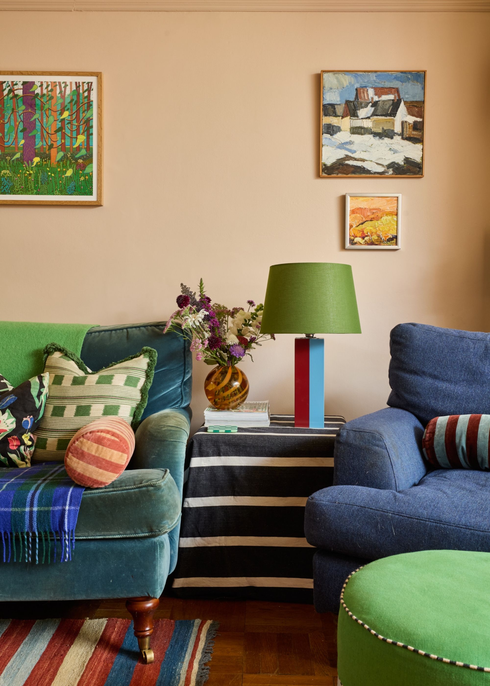 A living room with plaster pink walls, a teal sofa, a navy blue armchair, and pops of green decor, including a lamp, artwork, and cushions.