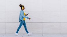 A woman wearing headphones and looking at her phone walks along a tiled area. 