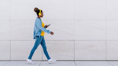 A woman wearing headphones and looking at her phone walks along a tiled area.