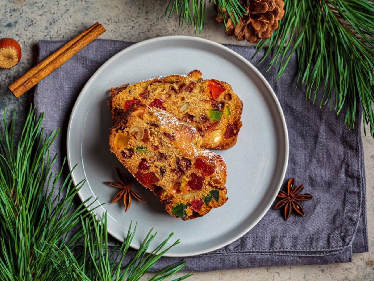 Two slices of fruitcake on a plate surrounded by pine branches, star anise, and cinnamon sticks