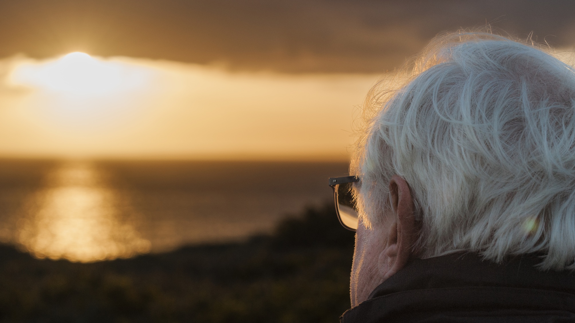 An older man looks toward a sunset over water.