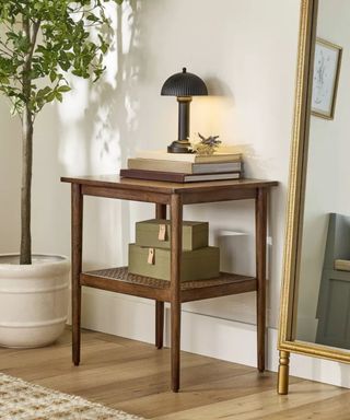 a small wooden side table sat in a hallway next to a large gold mirror styled with a stack of books and small black portable lamp