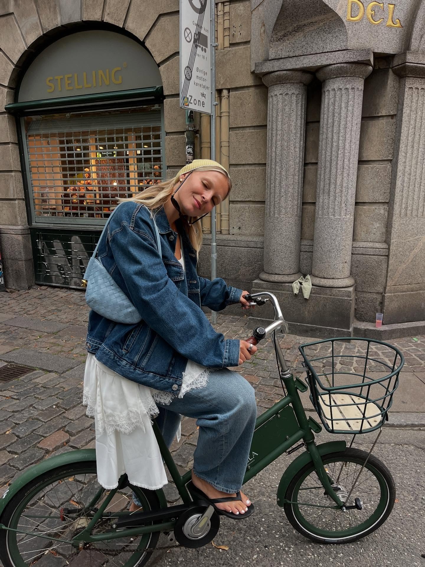 Hollie Mercedes wears the best autumn jacket in denim. She pairs it with jeans, a lace detail dress, a blue bag, and a yellow headscarf. She is on a bike, smiling at the camera in a recent image from Copenhagen Fashion Week, taken from her Instagram.