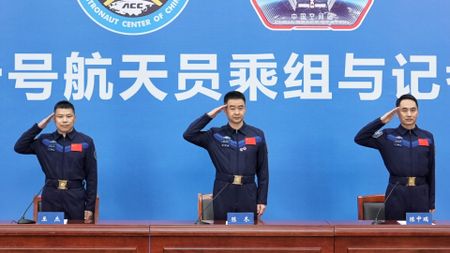 three men in blue flight suits salute behind a wooden desk covered in microphones