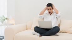 A young man looks shocked as he stares on his laptop while sitting on his sofa.