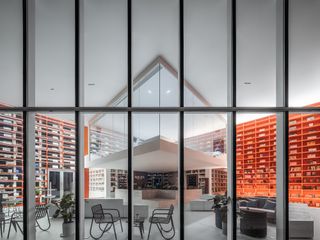 Looking into the interior through framed window, white room with orange shelving around the walls, black chairs and tables, high white ceiling with suspended glass viewing area, counter worktop, planters, glass double door on the left wall