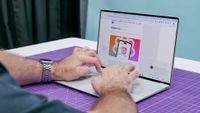 A person types on a laptop with a purple mat underneath. The screen displays an article with an image of a smartphone and colorful graphics.