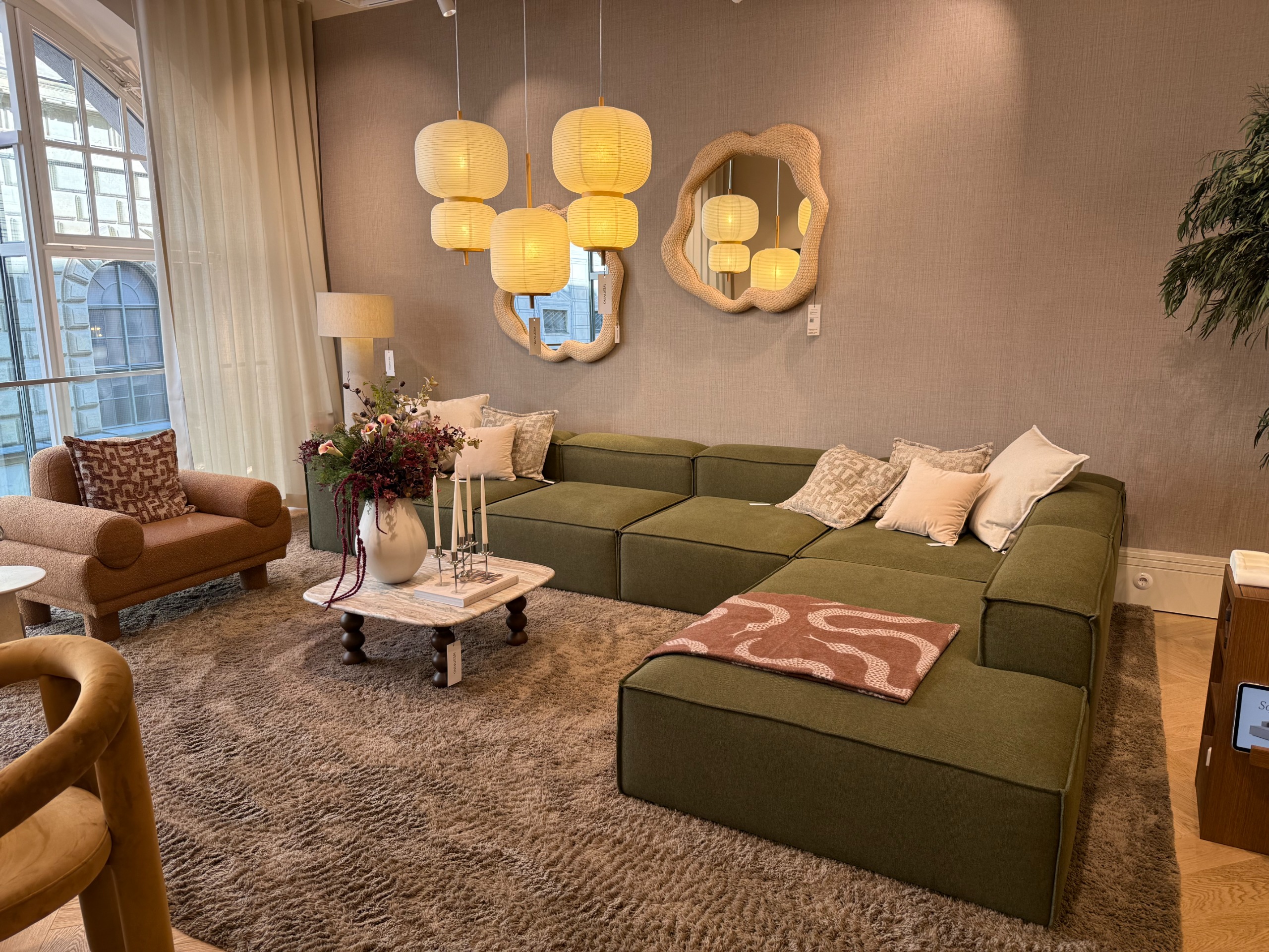 A living room setup in a showroom with a large sectional green sofa with neutral scatter cushions on the corners and a patterned throw on the closest edge, with mirrors on the walls and lighting above