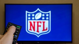 A hand holds a remote in front of a TV with the NFL logo on it