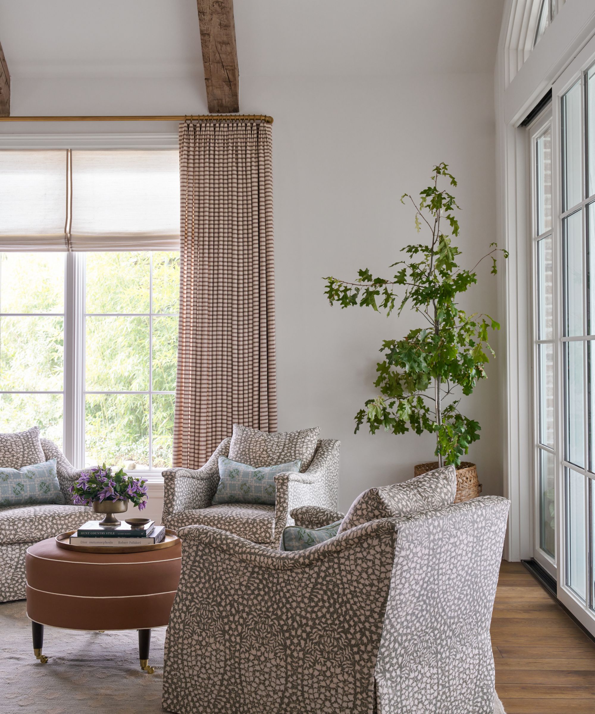 A transitional living room with white walls, large sliding glass doors leading to the garden on two walls, striped curtains, three matching patterned armchairs, and a large houseplant in the corner.