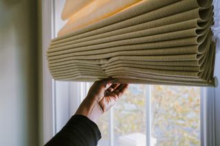 A hand pulling down beige blinds in front of a window.