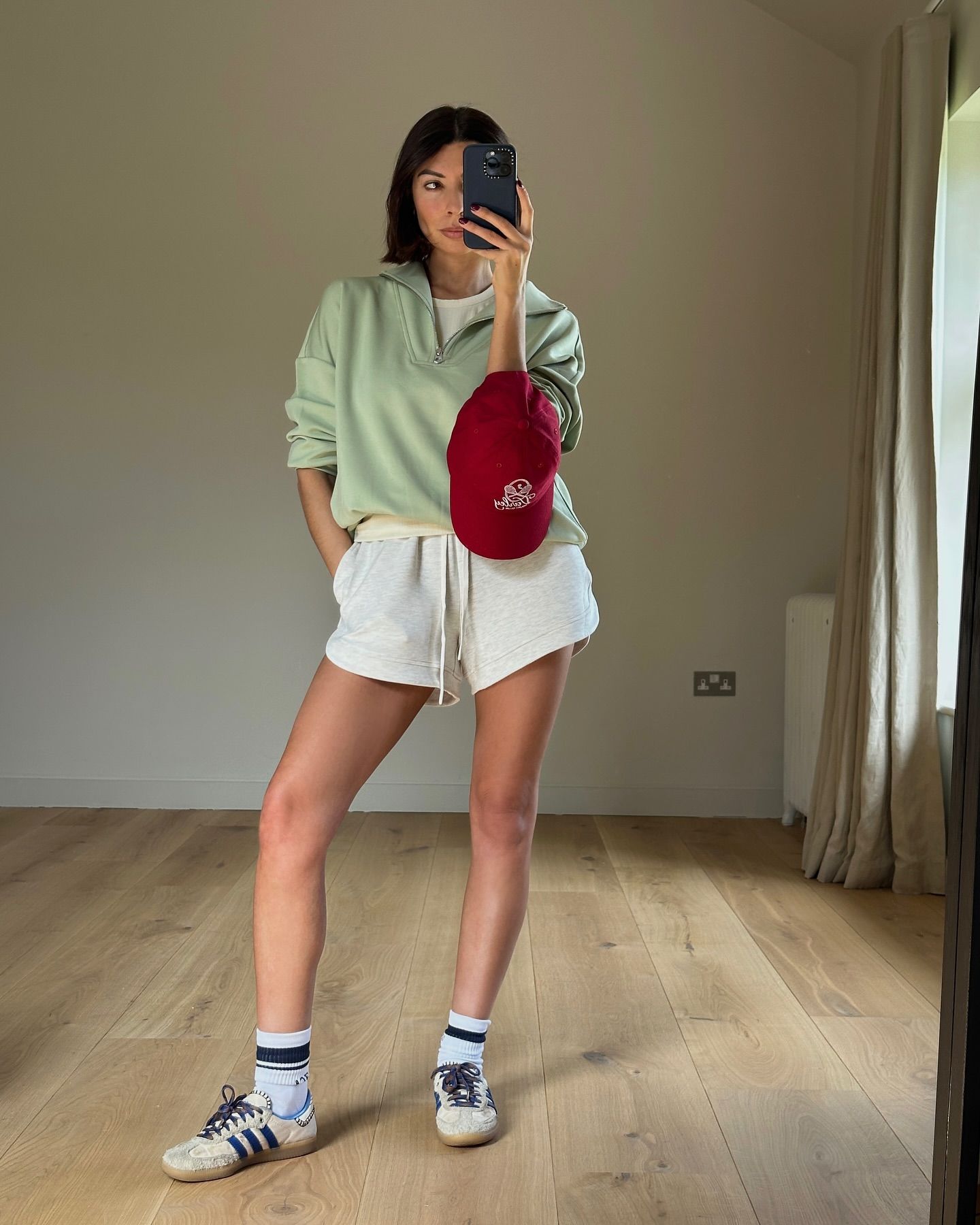 @smythsisters wearing mint green sweatshirt, white shorts, and has a maroon hat around her wrist. She is wearing black and white socks and the cream and blue Adidas x Wales Bonner collab trainer. She is taking a mirror selfie and is stood on light wooden floor in front of a white wall in a recent image from her Instagram.